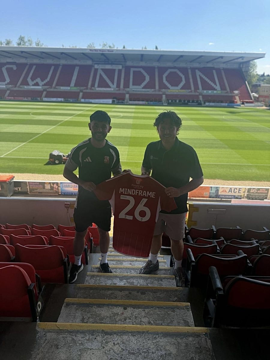 Jake Brown and Adam Labelle holding a Mindframe shirt in front of the SWINDON stand at the County Ground, celebrating Swindon Town's sport psychology partnership